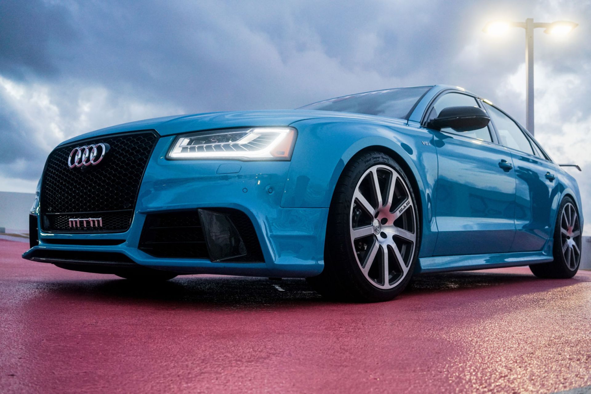 Blauer Audi auf nassem Asphalt bei bewölktem Himmel, fotografiert aus niedriger Perspektive mit eingeschaltetem Tagfahrlicht.
