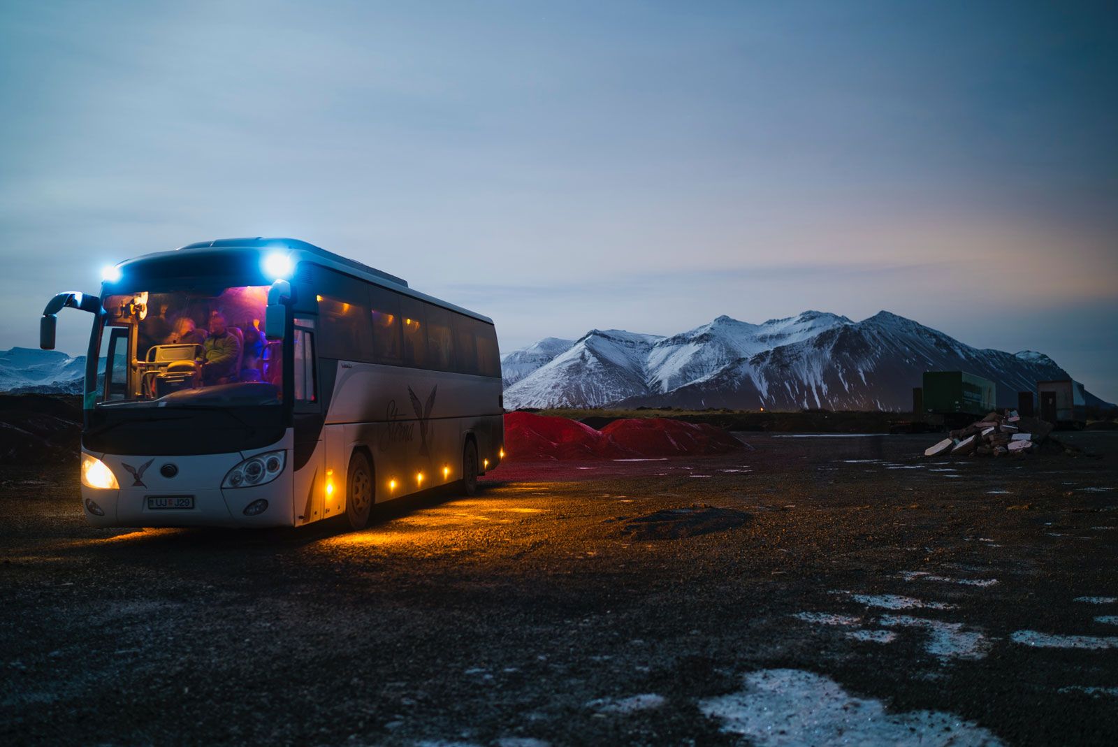 Beleuchteter Reisebus bei Dämmerung vor schneebedeckten Bergen auf einem weiten, teils verschneiten Schotterplatz.