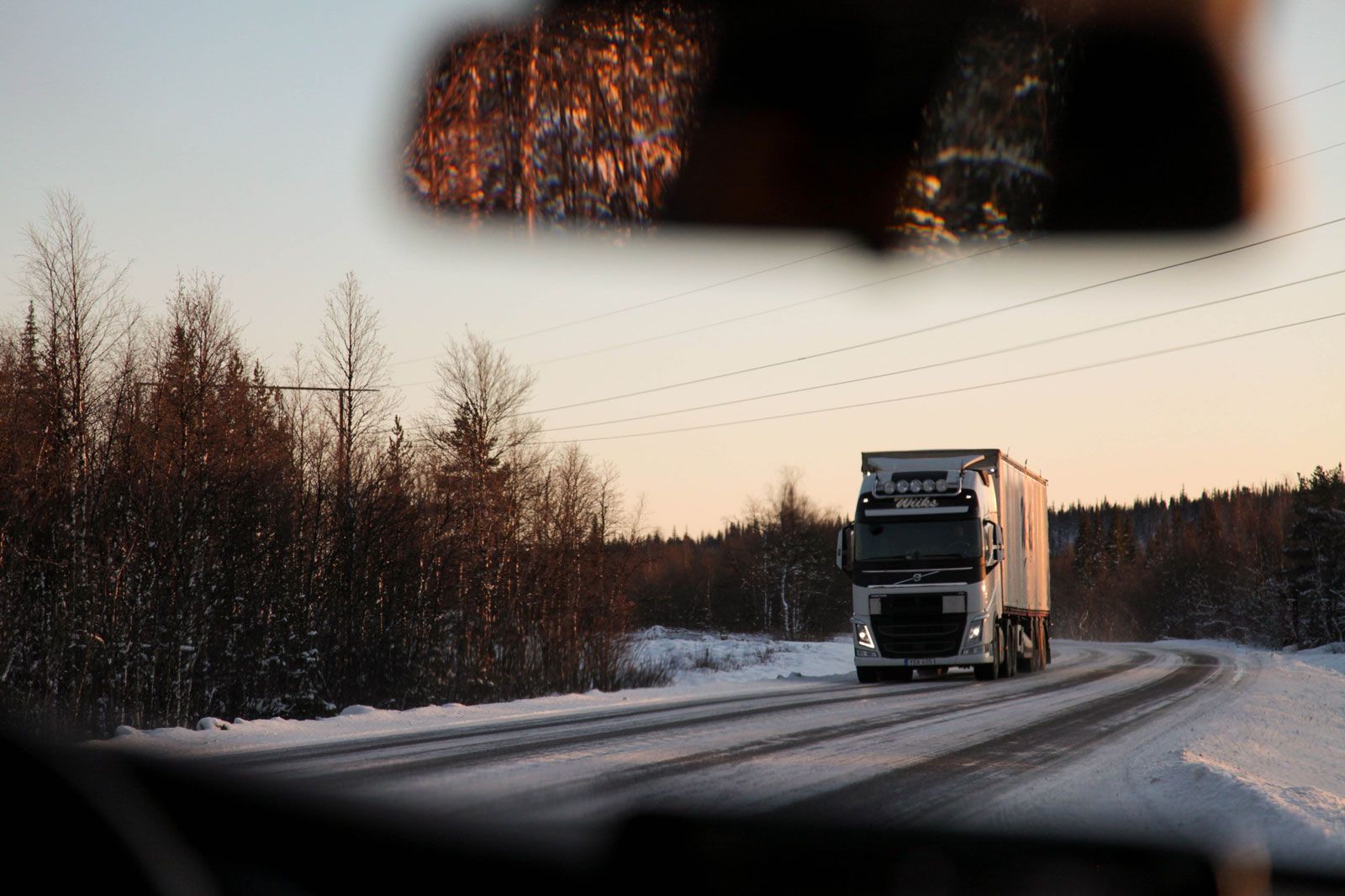LKW fährt auf schneebedeckter Landstraße durch winterliche Waldlandschaft, fotografiert durch die Frontscheibe eines Autos.